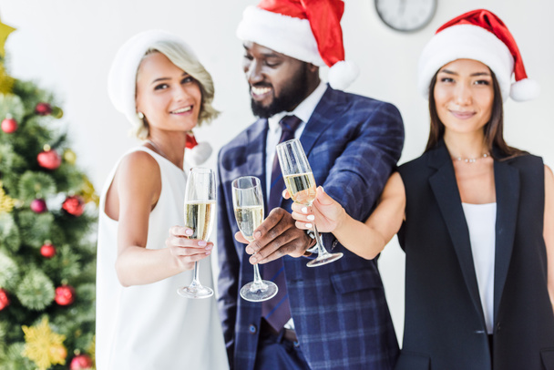 selective focus of multiethnic businesspeople in santa hats clinking with glasses of champagne in office - Foto, imagen