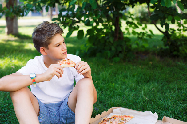 aranyos, fiatal fiú fehér póló, eszik pizzát, és mosolyog, már szórakozás a nyári park. - Fotó, kép