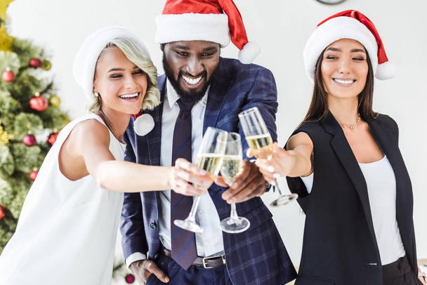 smiling multicultural businesspeople in santa hats clinking with glasses of champagne in office - Photo, Image