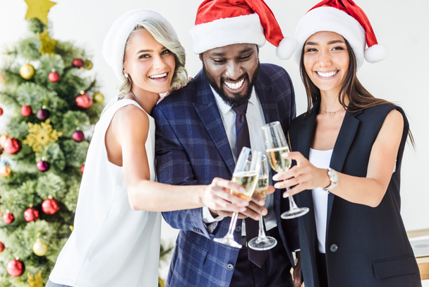 happy multiethnic businesspeople in santa hats clinking with glasses of champagne in office - Fotoğraf, Görsel