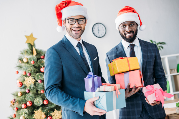 smiling multicultural businessmen in santa hats holding gift boxes in office - Fotó, kép