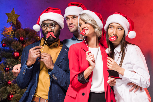 multicultural businesspeople holding lips and glasses on sticks at new year corporate party and looking away - Photo, Image
