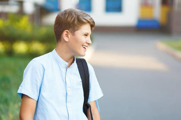 cute, young schoolboy in blue shirt with backpack  stands in front of his school. Back to school, education concept.  - Foto, Bild