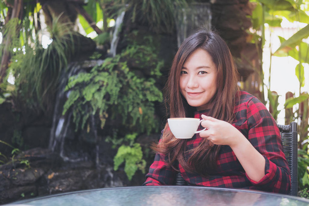 Портрет крупным планом: красивая азиатка держит белую кружку и пьет горячий кофе, чувствуя себя счастливой на зеленом фоне природы и водопада
 - Фото, изображение