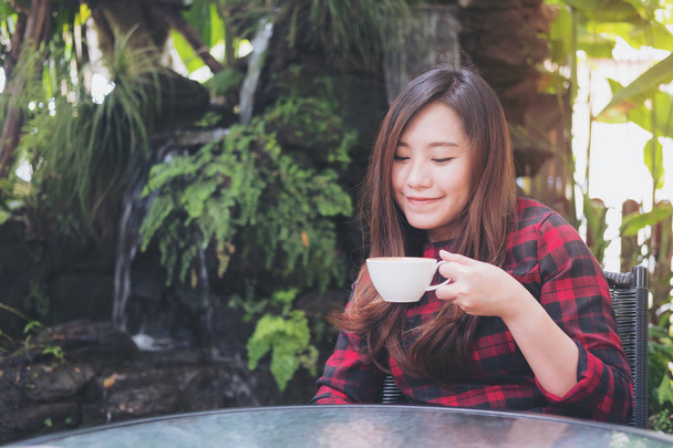 白いマグカップを押し、緑の自然と滝の庭の背景に幸せな気持ちでホット コーヒーを飲む美しいアジア女性のクローズ アップの肖像画のイメージ - 写真・画像