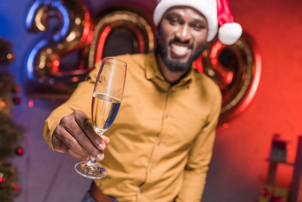 счастливый африканский американский бизнесмен в шляпе Санты показывает бокал шампанского на новогодней корпоративной вечеринке
 - Фото, изображение