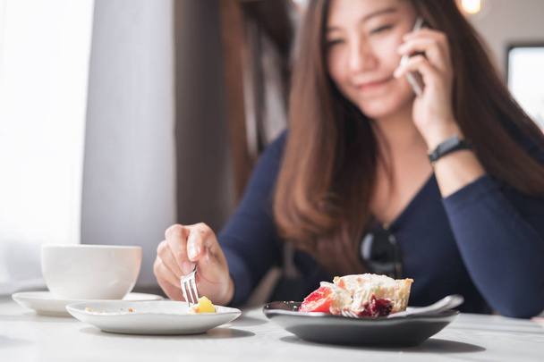 Closeup blur image of a smiley beautiful Asian woman holding and talking on smart phone with white coffee cup and dessert plates on wooden table in vintage cafe - Photo, Image