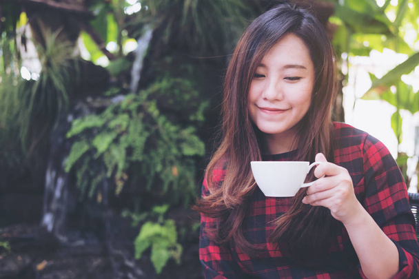 白いマグカップを押し、緑の自然と滝の庭の背景に幸せな気持ちでホット コーヒーを飲む美しいアジア女性のクローズ アップの肖像画のイメージ - 写真・画像