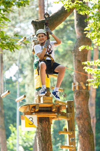 cute, sporty boy in white t shirt in the adventure rope activity park with helmet and safety equipment.Young boy playing and having fun doing activities outdoors. Hobby, active lifestyle concept - Фото, изображение