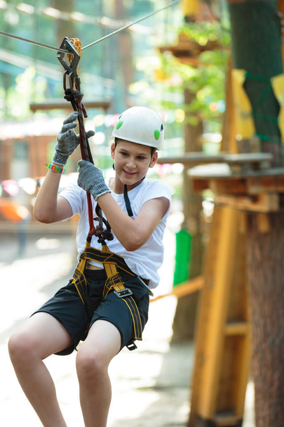 cute, sporty boy in white t shirt in the adventure rope activity park with helmet and safety equipment.Young boy playing and having fun doing activities outdoors. Hobby, active lifestyle concept - Фото, изображение