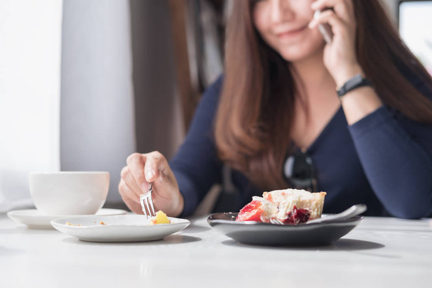 Крупным планом изображение улыбающейся красивой азиатки, держащей в руках и разговаривающей по смартфону с чашкой белого кофе и десертными тарелками на деревянном столе в винтажном кафе
 - Фото, изображение