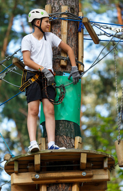 cute, sporty boy in white t shirt in the adventure rope activity park with helmet and safety equipment.Young boy playing and having fun doing activities outdoors. Hobby, active lifestyle concept - Foto, afbeelding