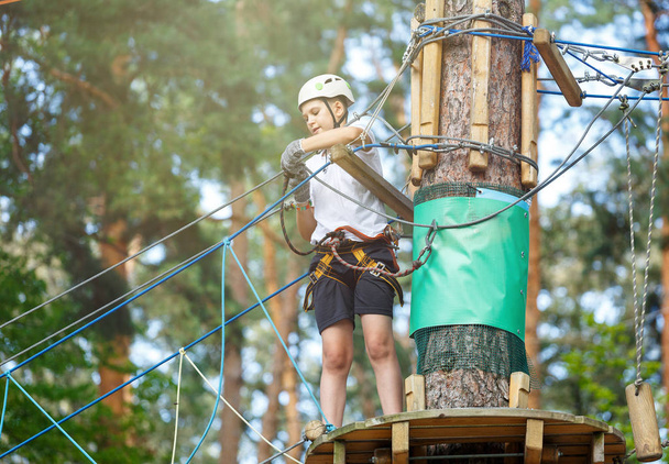 cute, sporty boy in white t shirt in the adventure rope activity park with helmet and safety equipment.Young boy playing and having fun doing activities outdoors. Hobby, active lifestyle concept - Foto, Bild