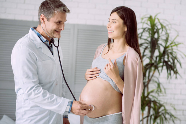 I feel the heartbeat. Happy positive nice doctor wearing stethoscope and smiling while feeling the heartbeat of a pregnant womans belly - Foto, immagini