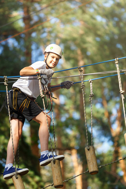 cute, sporty boy in white t shirt in the adventure rope activity park with helmet and safety equipment.Young boy playing and having fun doing activities outdoors. Hobby, active lifestyle concept - 写真・画像