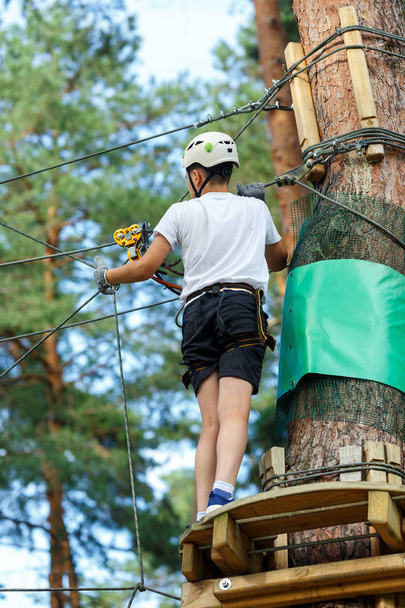 cute, sporty boy in white t shirt in the adventure rope activity park with helmet and safety equipment.Young boy playing and having fun doing activities outdoors. Hobby, active lifestyle concept - Foto, Imagem