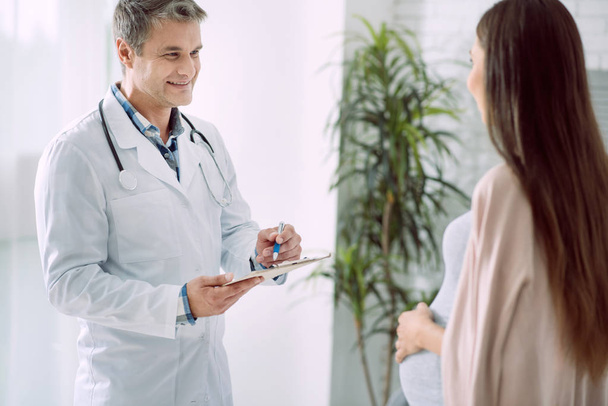 Absolutely healthy. Delighted positive professional doctor holding notes and looking at his patient while writing medical conclusion - Foto, imagen