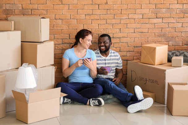 Interracial couple with cups of tea resting on floor near boxes in room. Moving into new house - Photo, image