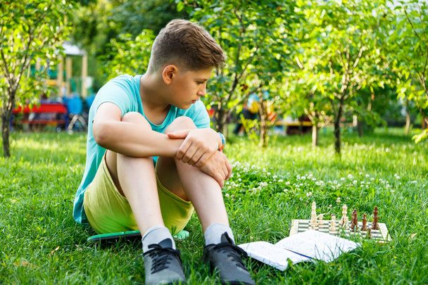 cute young boy in green t shirt plays chess on the chessboard in the park sitting on the grass. Education, lesson, training concept - Foto, immagini