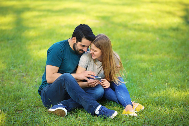 Heureux jeune couple avec téléphone portable assis sur l'herbe verte dans le parc
 - Photo, image