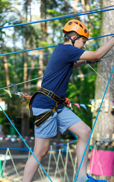Happy, cute, young boy in blue t shirt and orange helmet having fun and playing at adventure park, holding ropes and climbing wooden stairs. active lifestyle concept - Photo, Image