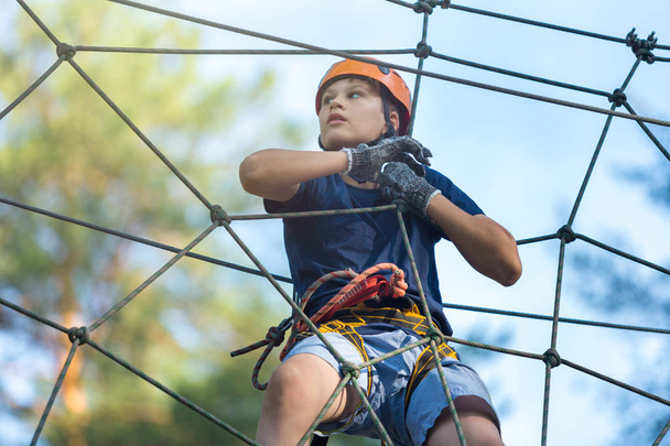 Happy, cute, young boy in blue t shirt and orange helmet having fun and playing at adventure park, holding ropes and climbing wooden stairs. active lifestyle concept - Zdjęcie, obraz