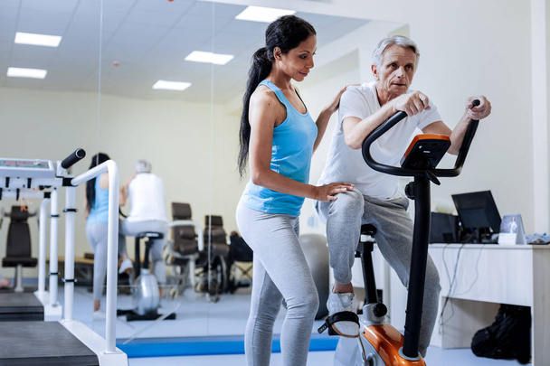 Exhausted aged man sitting on the exercise bike while his trainer smiling - Foto, Imagen