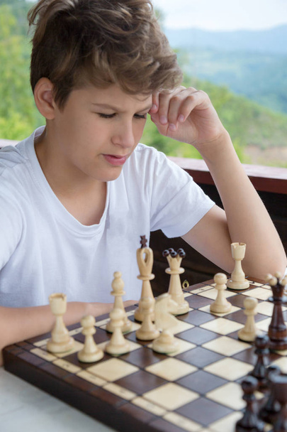cute, smart, 11 years old boy in white shirt sits in the classroom and plays chess on the chessboard. Training, lesson, hobby, education concept. intellectual game - Zdjęcie, obraz
