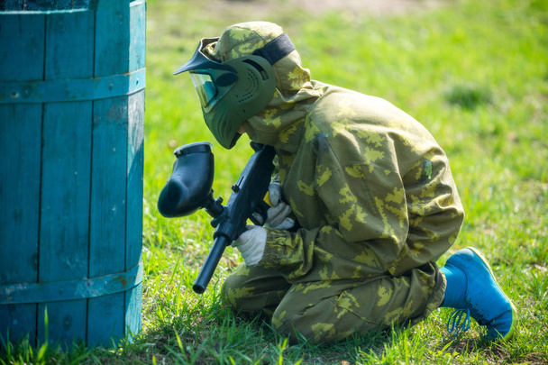Jongen in de camouflage houdt een paintball pistool in één hand en beschermende helm, staande op het veld. sport levensstijl concept  - Foto, afbeelding