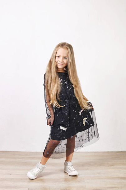 Little blonde girl with long golden hair wearing black dress with stars & planets, dancing, smling & having fun, white wall background. Five years old blonde female child posing. Copy space, close up. - Foto, Imagem