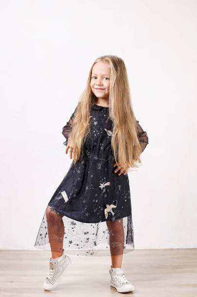 Little blonde girl with long golden hair wearing black dress with stars & planets, dancing, smling & having fun, white wall background. Five years old blonde female child posing. Copy space, close up. - Zdjęcie, obraz