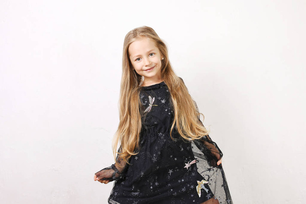 Little blonde girl with long golden hair wearing black dress with stars & planets, dancing, smling & having fun, white wall background. Five years old blonde female child posing. Copy space, close up. - Foto, Bild
