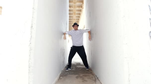 Een jonge kerel in een hoed dansen knallen tussen twee witte muren aan de buitenkant in de zomer - Video