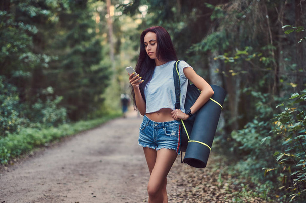 Charming hipster girl dressed in a shirt and shorts with tourist mat and backpack using a smartphone while standing in a summer forest. - Photo, Image