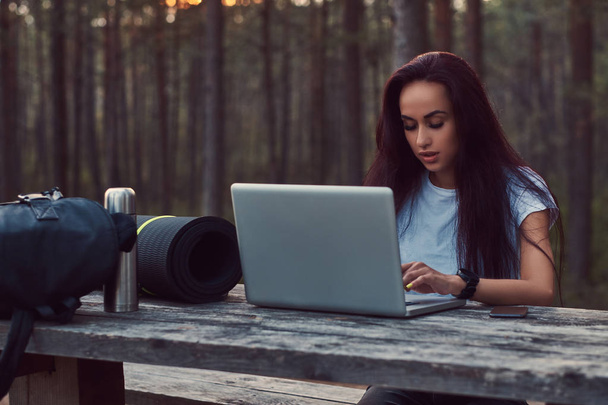 Turista chica hipster en camisa blanca trabajando en un ordenador portátil mientras está sentado en un banco de madera en el hermoso bosque de otoño
. - Foto, imagen