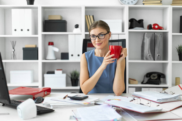 Una joven está sentada en una mesa en la oficina, sosteniendo una taza roja en la mano y trabajando con documentos
. - Foto, imagen
