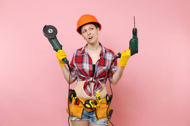 Ενέργεια διασκέδαση πολυτεχνίτης γυναίκα στα γάντια, προστατευτικό κράνος πορτοκαλί, κιτ εργαλείων ζώνη γεμάτη μέσα κρατώντας μηχανικό πριόνι ηλεκτρικό τρυπάνι που απομονώνονται σε ροζ φόντο. Θηλυκό σε αρσενικό το έργο. Ανακαίνισης - Φωτογραφία, εικόνα