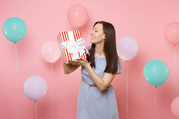 Portrait of joyful attractive woman in blue dress looking on red box with gift present on pastel pink background with colorful air balloons. Birthday holiday party, people sincere emotions concept - Foto, afbeelding