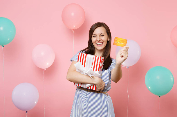 Portrait of beautiful smiling young woman in blue dress holding credit card and red box with gift present on pink background with colorful air balloons. Birthday holiday party, people sincere emotion - Photo, Image