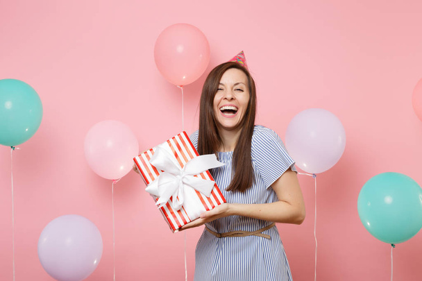 Portrait of laughing pretty woman in birthday hat and blue dress holding red box with gift present on pastel pink background with colorful air baloons. Birthday holiday party, people sincere emotions - Zdjęcie, obraz
