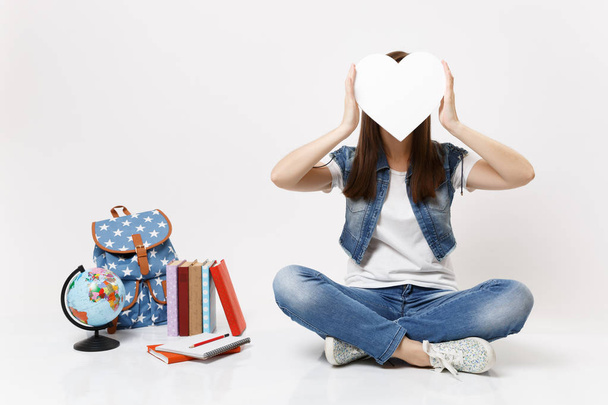 Young brunette woman student covering face with white heart with copy space sitting near globe backpack, school books isolated on white background. Education in high school university college concept - Photo, Image