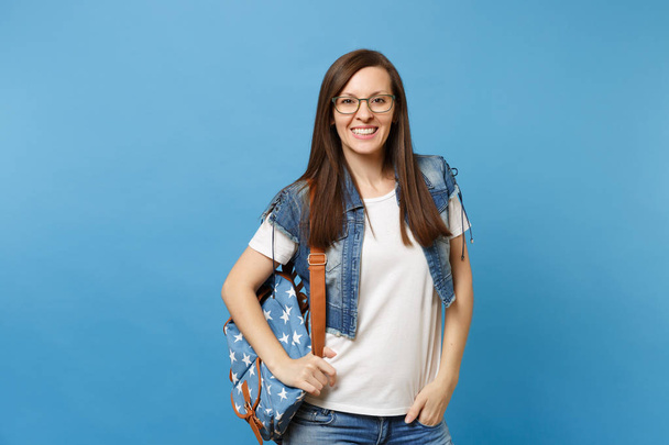 Young casual smart woman student in white t-shirt, denim clothes with backpack wearing glasses keeping hand in pocket isolated on blue background. Education in high school university college concept - Photo, image