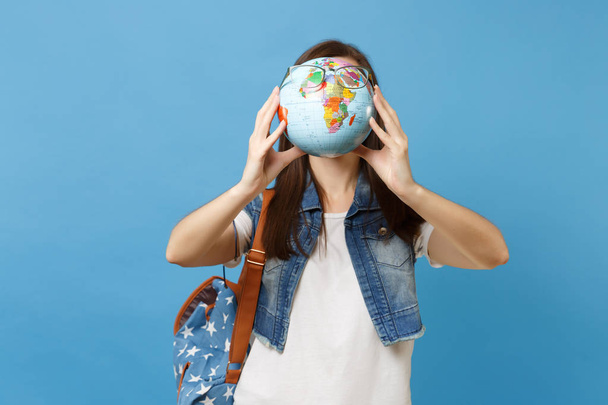 Retrato de jovem estudante morena casual com mochila segurando e cobrindo rosto com globo mundial com óculos isolados em fundo azul. Educação no ensino médio conceito faculdade universitária
 - Foto, Imagem