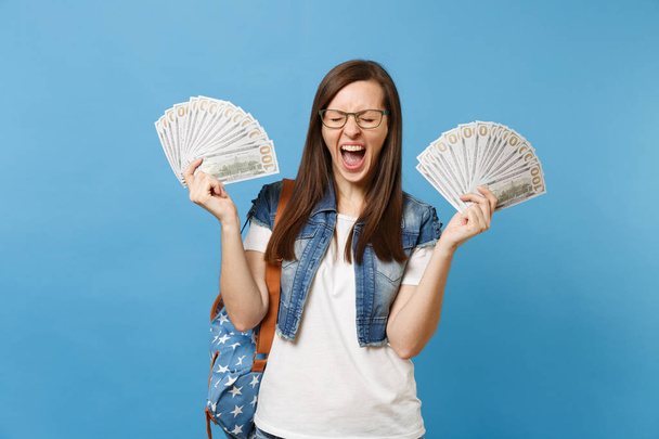 Young crazy woman student in glasses with backpack with closed eyes screaming hold bundle lots of dollars, cash money isolated on blue background. Education in high school university college concept - Photo, Image
