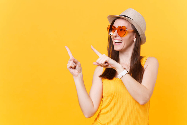 Portrait de jeune femme souriante en chapeau d'été en paille, lunettes orange pointant index de côté espace de copie isolé sur fond jaune. Les gens émotions sincères, concept de style de vie. Espace publicitaire
 - Photo, image