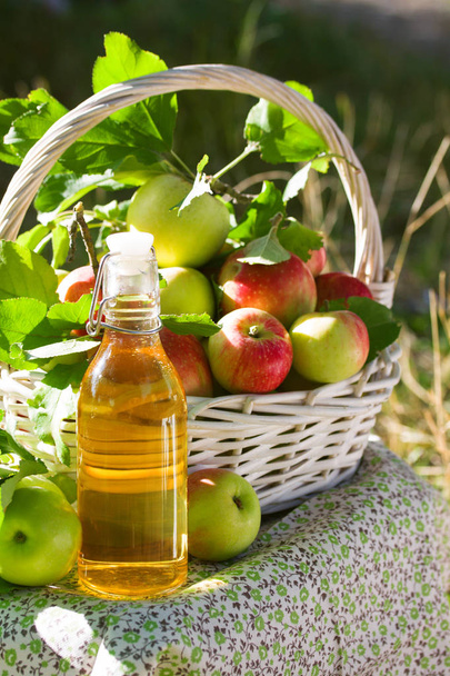White wicker basket with green and red apples, apple cider, juice or vinegar in glass bottle, leaves on nature background, sunlight. Summer, autumn, fresh apple harvest, harvesting. - Фото, зображення