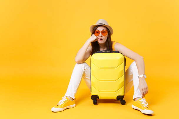Tylsää turisti nainen kesällä rento vaatteet hattu istua matkalaukku, odota kone eristetty keltainen oranssi tausta. Tyttö matkustaa ulkomaille viikonloppumatkalle. Ilmalennon käsite
 - Valokuva, kuva