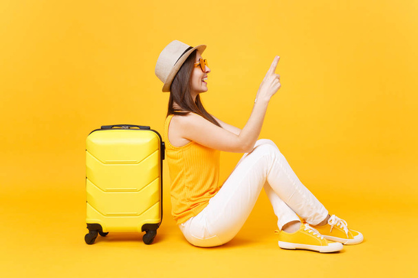 旅行観光女性夏カジュアルな服を着て、黄色オレンジ色の背景に分離されたスーツケースと帽子座っています。女性の乗客は、週末の息抜きに旅行に海外旅行します。空気の旅のコンセプト - 写真・画像