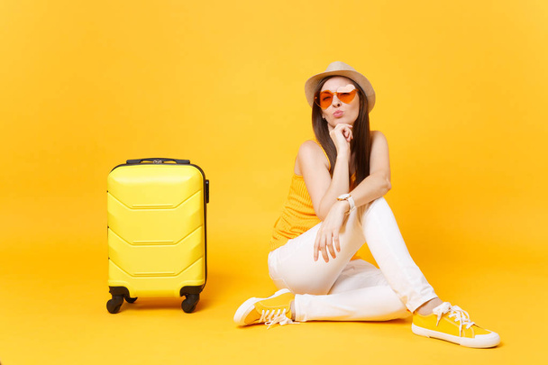 旅行観光女性夏カジュアルな服を着て、黄色オレンジ色の背景に分離されたスーツケースと帽子座っています。女性の乗客は、週末の息抜きに旅行に海外旅行します。空気の旅のコンセプト - 写真・画像