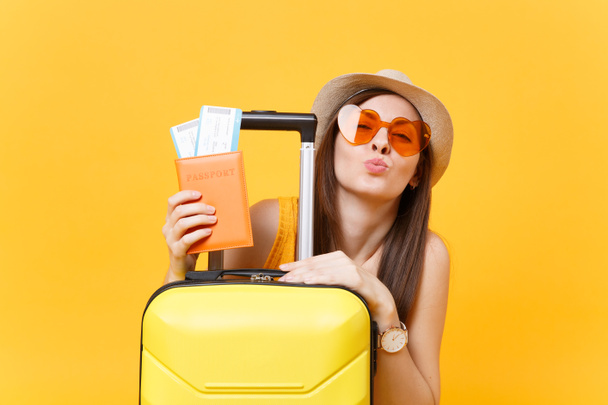 Весела туристична жінка в літньому повсякденному одязі, капелюсі, помаранчевих сонцезахисних окулярах з валізою, паспорт ізольований на жовтому тлі. Пасажири, які виїжджають за кордон у відпустку у вихідні дні. Концепція авіарейсу
 - Фото, зображення
